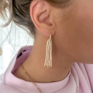 Könnte beinhalten: Goldfarbene Ohrringe mit einem rechteckigen Pfosten und mehreren Strängen kleiner Perlen, die herunterhängen. Eine goldene Halskette ist ebenfalls sichtbar. Die Ohrringe werden an einem Ohr getragen, vor einem rosa Hintergrund.