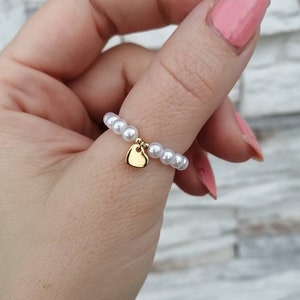 May include: A white pearl ring with a gold heart charm. The ring is worn on a finger.