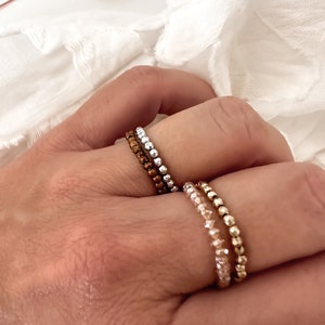 May include: Three beaded rings stacked on a finger. The top ring is silver, the middle ring is copper, and the bottom ring is gold.