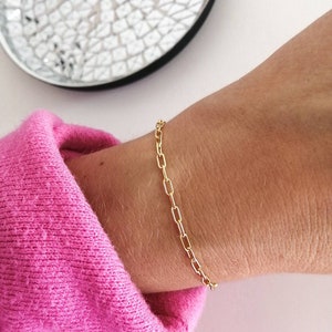 Puede incluir: Un brazalete de cadena dorada con un delicado diseño de cadena de eslabones cuadrados. El brazalete se muestra en la muñeca de una persona.