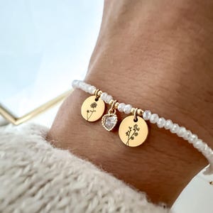 May include: A gold bracelet with white beads and three charms. Two round charms have floral designs, and a heart-shaped charm sparkles with clear stones. The bracelet is worn on a wrist.