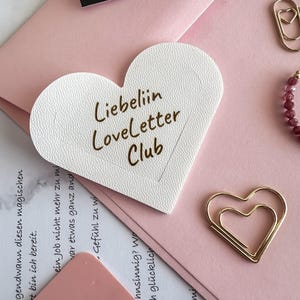 Könnte beinhalten: Eine weiße, texturierte Karte in Herzform mit dem braunen Aufdruck "Liebeliin Loveletter Club" auf der Vorderseite. Die Karte liegt neben einer goldenen herzförmigen Büroklammer und einem rosa Umschlag, mit bedrucktem Papier darunter.
