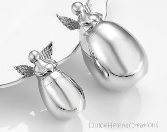 May include: Two silver angel-shaped cremation urns. The urns are egg-shaped and have a small angel figure on top holding a heart.