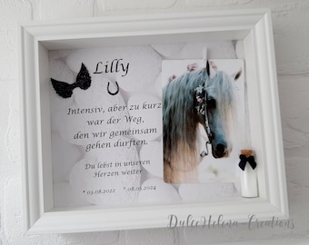 May include: A white picture frame with a black and white photo of a horse with a flower crown. The frame has a black bow, a horseshoe, and a small glass bottle with a black bow. The text "Lilly" is written in black at the top of the frame. The text "Intensiv, aber zu kurz war der Weg, den wir gemeinsam gehen durften. Du lebst in unseren Herzen weiter * 03.08.2022 + 08.08.2024" is written in black below the photo. The text "Dulce Helena-Creations" is written in black at the bottom of the frame.