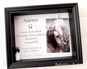 May include: A black framed picture with a white background and a photo of a white horse. The text "Adonis" is at the top of the frame with a horseshoe symbol. The text "Wenn Liebe allein Dich hätte retten können, Du hättest ewig gelebt." is below the photo. The text "Du bist für immer in unseren Herzen." is below the text. The dates "*14.07.2005" and "1 25.09.2022" are below the text. The text "DulceHelena Creations" is at the bottom of the frame.