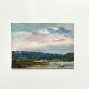 Puede incluir: Una pintura al óleo original que representa una escena de paisaje. La obra de arte presenta una masa de agua en primer plano, una línea de árboles verde oscuro y azul, y un cielo nublado con tonos rosas y azules. La pintura está firmada en la esquina inferior izquierda.