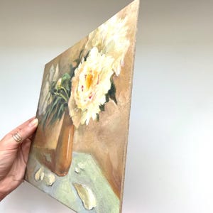 May include: A square canvas painting depicting a still life of white and yellow peonies in a brown vase. The flowers are arranged on a light green surface against a tan background. Several white flower petals are scattered on the surface.