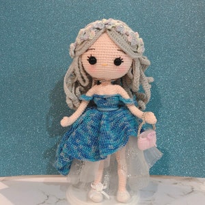 May include: A crocheted doll wearing a blue and white dress with a white tulle skirt and a flower crown. The doll is holding a pink purse.