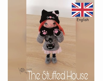 May include: A crocheted doll wearing a black cat hat, a gray sweater with a black cat design, and black shoes. The doll has red hair and pink cheeks. The doll is standing on a wooden surface. The text "The Stuffed House" is visible in the image.