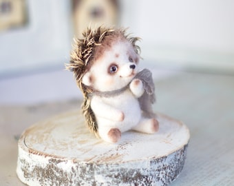 May include: A small, white, needle-felted hedgehog with brown quills and a gray scarf sits on a wooden log. The hedgehog has black eyes and a pink nose.