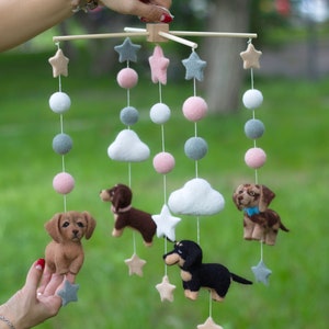 May include: A felt baby mobile with a wooden frame, featuring four dachshund dogs, clouds, and stars. The dogs are brown, black, and tan. The clouds are white. The stars are yellow. The mobile is hanging from a wooden rod.