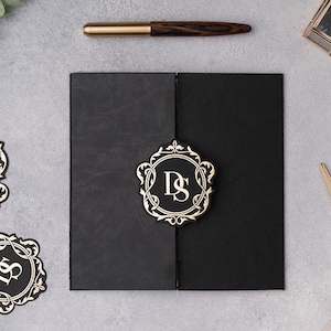 Può includere: Invito di nozze nero con un emblema dorato con le iniziali "DS". L'invito è abbinato a adesivi coordinati, una penna d'oro, forbici e un portagioie. È incluso anche un nastro.