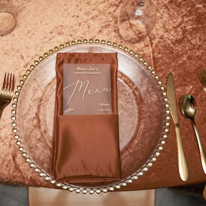 Puede incluir: Un plato de sitio de vidrio transparente con borde dorado de cuentas, que incluye una servilleta de satén color óxido doblada y un menú translúcido. El texto dice Nurse Jen's Graduation Celebration, Menu y Entrees Lamb Chops.