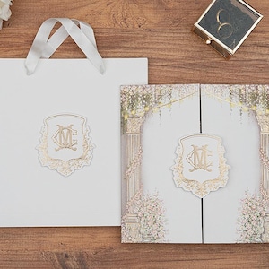 Könnte beinhalten: Eine weiße Geschenktüte und ein Einladungsset mit goldenen Akzenten sind auf einer Holzoberfläche angeordnet. Die Einladung zeigt ein florales Design und ein goldenes Emblem. Ein goldener Stift und eine Ringbox sind ebenfalls vorhanden.