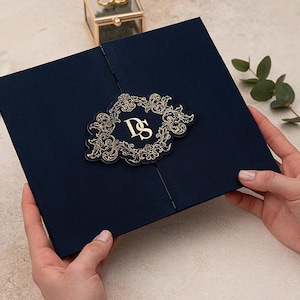 Op de afbeelding: Marineblauwe uitnodiging met een gouden filigraan ontwerp en de initialen "DS". De uitnodiging wordt door handen vastgehouden en is omgeven door decoratieve elementen, waaronder bloemen en groen. Een huwelijksuitnodiging.