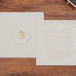 May include: A square cream-colored wedding invitation suite featuring gold foil lettering and a decorative border. The invitation displays the names Berrin & Gokhan with a matching envelope, a wax seal with a gold monogram, and a dark brown pen.