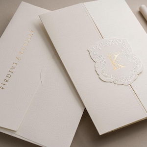 May include: Two off-white textured paper invitation folders. One folder features the gold foil text FIRDEVS & KUBILAY. The other folder is closed with a die-cut off-white seal featuring an embossed border and a gold foil letter K in the centre.