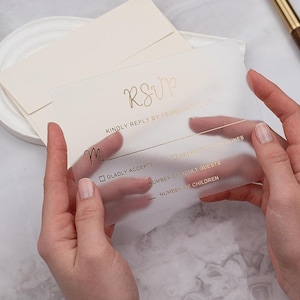 May include: A clear RSVP card with gold lettering is held between two hands. The card includes the word "RSVP" and prompts for a reply. A cream-colored envelope and a gold pen are in the background. Wedding stationery.