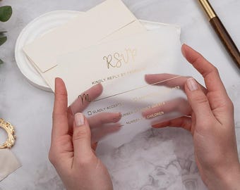 Op de afbeelding: Een transparante RSVP-kaart met gouden letters wordt vastgehouden. De kaart bevat het woord "RSVP" en vraagt om een antwoord. Een crème-kleurige envelop en een gouden pen staan op de achtergrond. Bruiloft briefpapier.