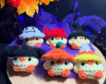 May include: A collection of six crocheted mushroom figures with various hat designs. The mushrooms are cream-colored with orange accents and black eyes. The hats are in different colors, including white, red, and black. The figures are displayed on a wooden surface.