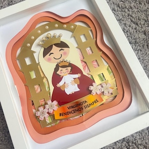 May include: A framed paper art piece featuring a layered design of a religious figure with a child, adorned with gold accents and a crown. The artwork includes the text "VIRGENCITA BENDICENOS SIEMPRE" and is set within a white frame.