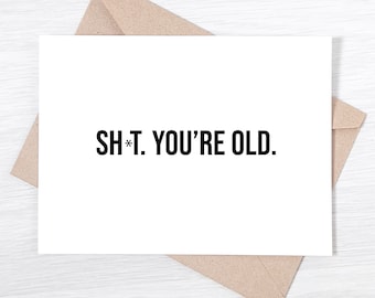 May include: A white greeting card with the text "SH*T. YOU'RE OLD." in black, placed on a brown envelope. The card and envelope are on a light-colored wooden surface. The card is a humorous birthday card.