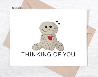 May include: A white card with a cartoon illustration of a brown voodoo doll with a red heart and the text "Thinking of You".