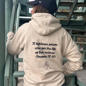 Könnte beinhalten: Ein beiger Hoodie mit einem schwarzen Aufdruck, der "Ein gerechter Mensch sorgt für das Leben seines Tieres -Sprüche 12:10" lautet und eine kleine Illustration von zwei ineinander verschlungenen Händen.
