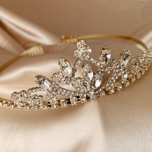 Pode incluir: Uma tiara dourada com strass transparentes e um design delicado. A tiara &eacute; feita de metal e tem um visual cl&aacute;ssico e elegante.