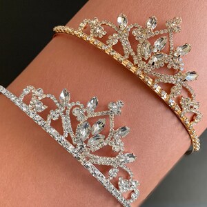 Pode incluir: Duas coroas de cabe&ccedil;a de ouro e prata com strass. As coroas est&atilde;o no bra&ccedil;o de uma pessoa.
