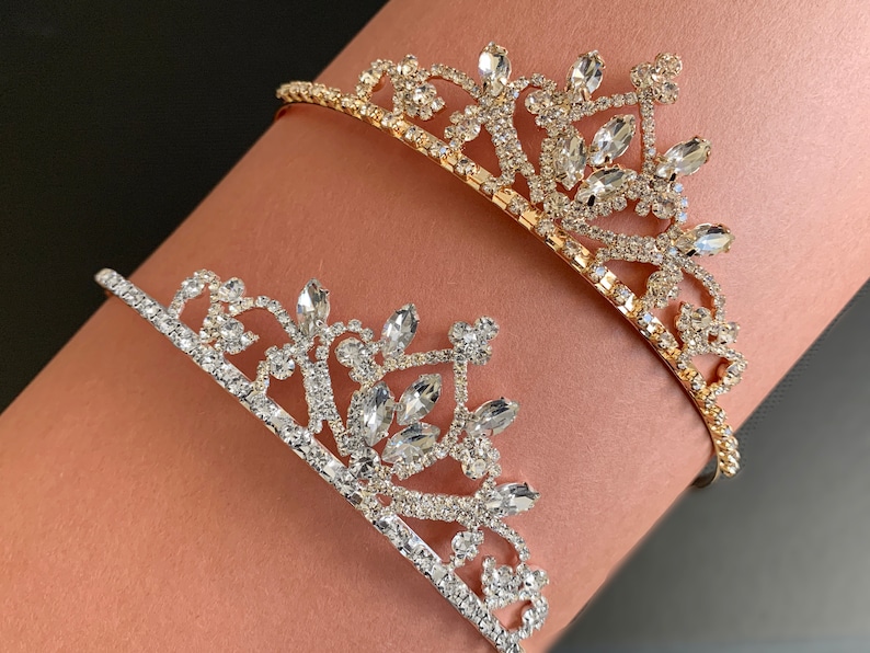 Pode incluir: Duas coroas de cabe&ccedil;a de ouro e prata com strass. As coroas est&atilde;o no bra&ccedil;o de uma pessoa.