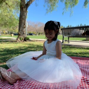 A 7-year-old girl is sitting on the grass wearing a white tulle flower girl dress, with a mini rhinestone tiara on her head. The tiara is densely adorned with sparkling rhinestones.