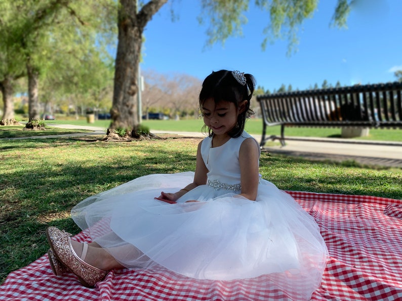 A 7-year-old girl is sitting on the grass wearing a white tulle flower girl dress, with a mini rhinestone tiara on her head. The tiara is densely adorned with sparkling rhinestones.