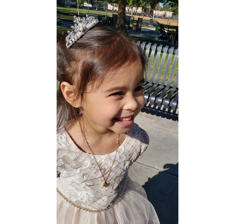 May include: A young girl wearing a white dress with a tulle skirt and a gold necklace with a heart pendant. She is smiling and wearing a sparkly tiara in her hair.