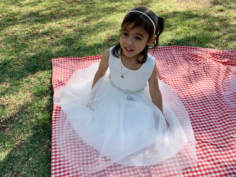 A young girl wearing a white sleeveless dress with a tulle skirt and a jeweled belt. She is sitting on a red and white checkered picnic blanket in a grassy area, with a gold frame headband with rhinestones and pearls.