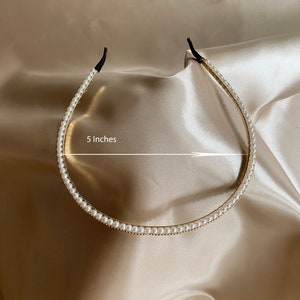 A gold-toned headband adorned with small, white faux pearls. The headband is approximately 5 inches in diameter and features black elastic ends for a comfortable fit. The accessory is displayed on a satin surface.