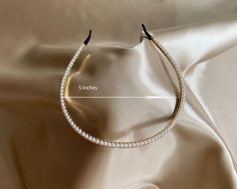 A gold-toned headband adorned with small, white faux pearls. The headband is approximately 5 inches in diameter and features black elastic ends for a comfortable fit. The accessory is displayed on a satin surface.