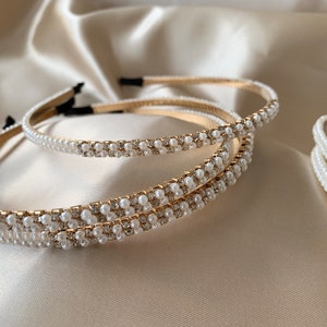 Several gold headbands with white faux pearls  and small clear rhinestones. The headbands are arranged on a satin surface. The headbands have a thin, gold band and are decorated with rows of pearls and rhinestones.
