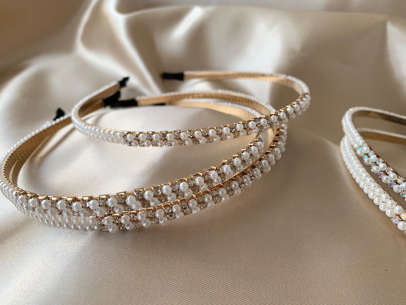 Several gold headbands with white faux pearls  and small clear rhinestones. The headbands are arranged on a satin surface. The headbands have a thin, gold band and are decorated with rows of pearls and rhinestones.