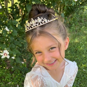 Pode incluir: Uma menina pequena usando um vestido branco de renda com uma tiara dourada adornada com strass transparentes. A menina est&aacute; sorrindo e olhando para a c&acirc;mera.