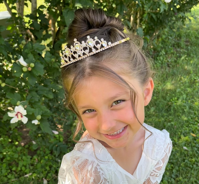 Pode incluir: Uma menina pequena usando um vestido branco de renda com uma tiara dourada adornada com strass transparentes. A menina est&aacute; sorrindo e olhando para a c&acirc;mera.