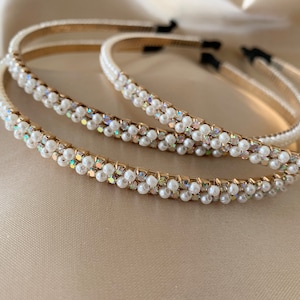 Close-up of several gold headbands adorned with rows of small, white pearls and iridescent rhinestones. The headbands have a thin, gold band and are arranged on a soft, cream-colored surface. The headbands are a fashion accessory.