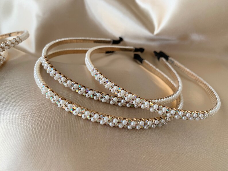 Several pearl-embellished headbands with gold-toned bands are displayed on a satin surface. The headbands feature rows of small, white pearls and iridescent accents, creating a delicate and elegant appearance. The headbands are a hair accessory.