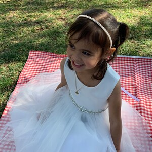 A young girl wearing a white dress with a tulle skirt and a pearl and rhinestone headband. The dress has a jeweled belt. She is sitting on a red and white checkered picnic blanket on a grassy area.