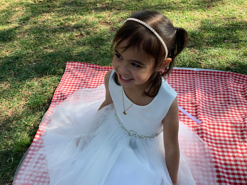 A young girl wearing a white dress with a tulle skirt and a pearl and rhinestone headband. The dress has a jeweled belt. She is sitting on a red and white checkered picnic blanket on a grassy area.