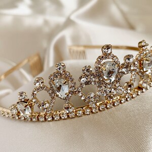 Pode incluir: Uma tiara dourada com strass transparentes e uma pedra central em forma de l&aacute;grima.