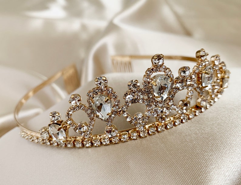 Pode incluir: Uma tiara dourada com strass transparentes e uma pedra central em forma de l&aacute;grima.