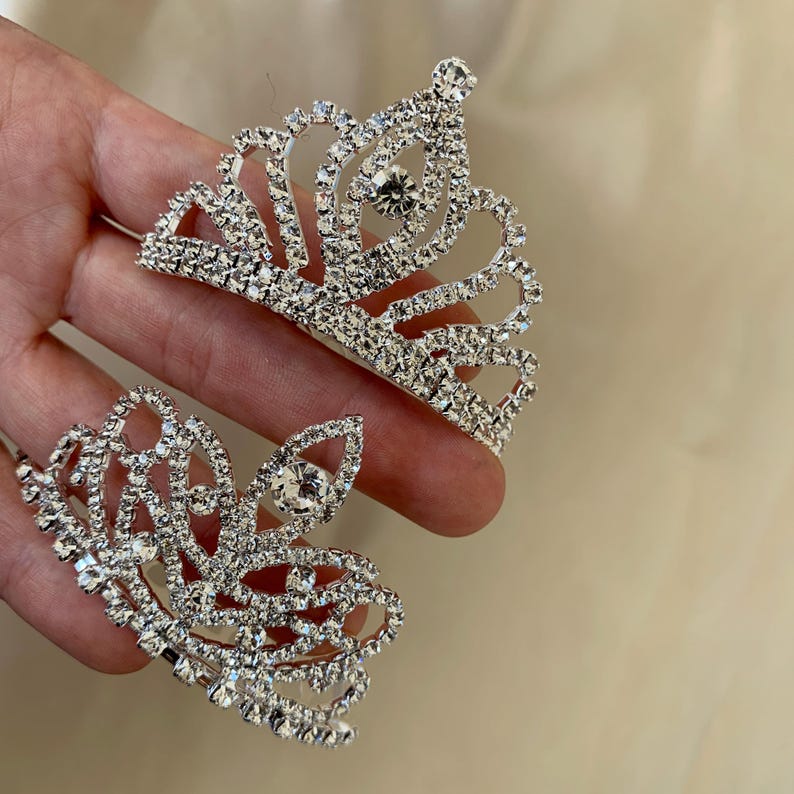Two mini tiaras are held in one hand—one with a rounded shape adorned with rhinestones, and the other with a slightly elongated shape, also set with rhinestones. Both are mini-sized and sparkle in the light against a beige background.