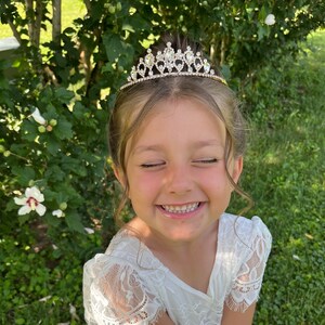 Pode incluir: Uma menina pequena usando um vestido branco de renda com uma tiara dourada adornada com strass transparentes. Ela est&aacute; sorrindo com os olhos fechados.
