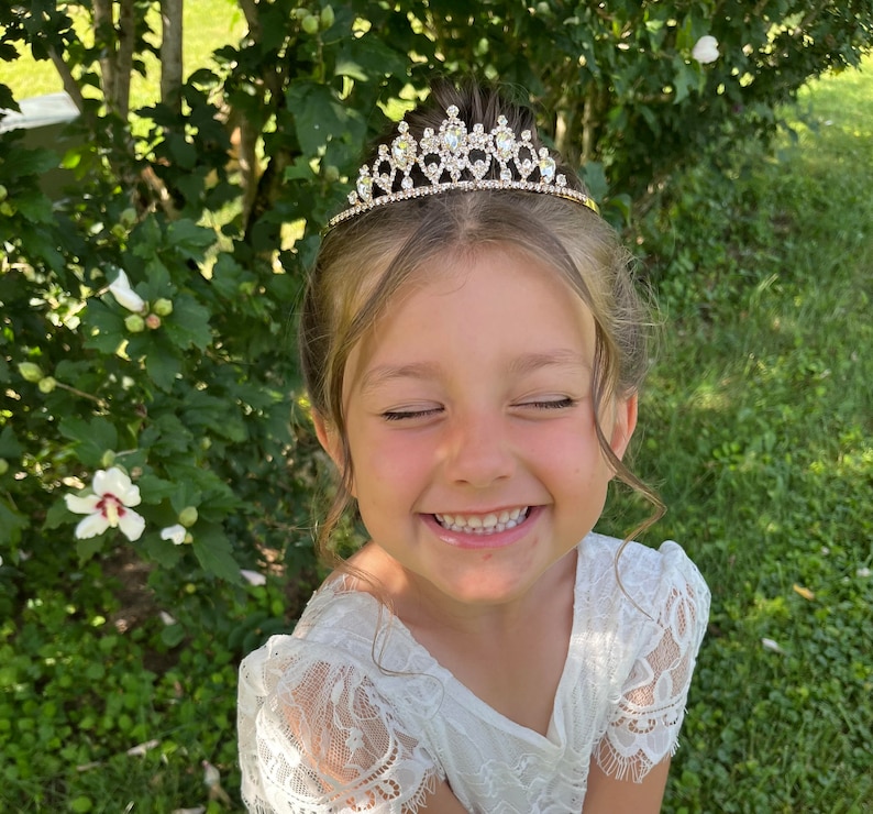 Pode incluir: Uma menina pequena usando um vestido branco de renda com uma tiara dourada adornada com strass transparentes. Ela est&aacute; sorrindo com os olhos fechados.